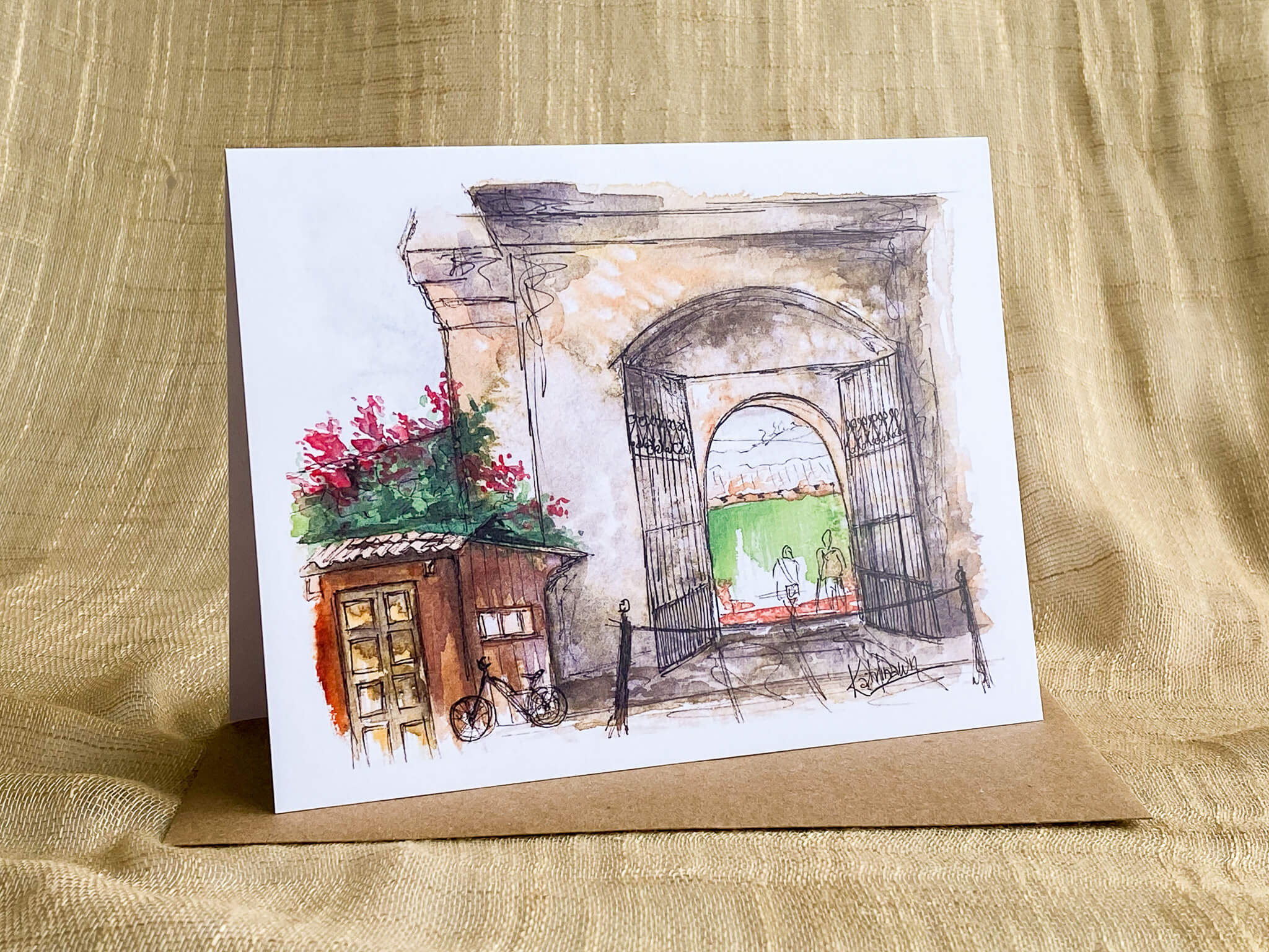 Watercolor sketch greeting card of the stone archway entrance to the San Francisco cathedral located in Antigua, Guatemala, with wrought iron gates, bougainvillea flowers, and a bicycle, shown with a kraft paper envelope.