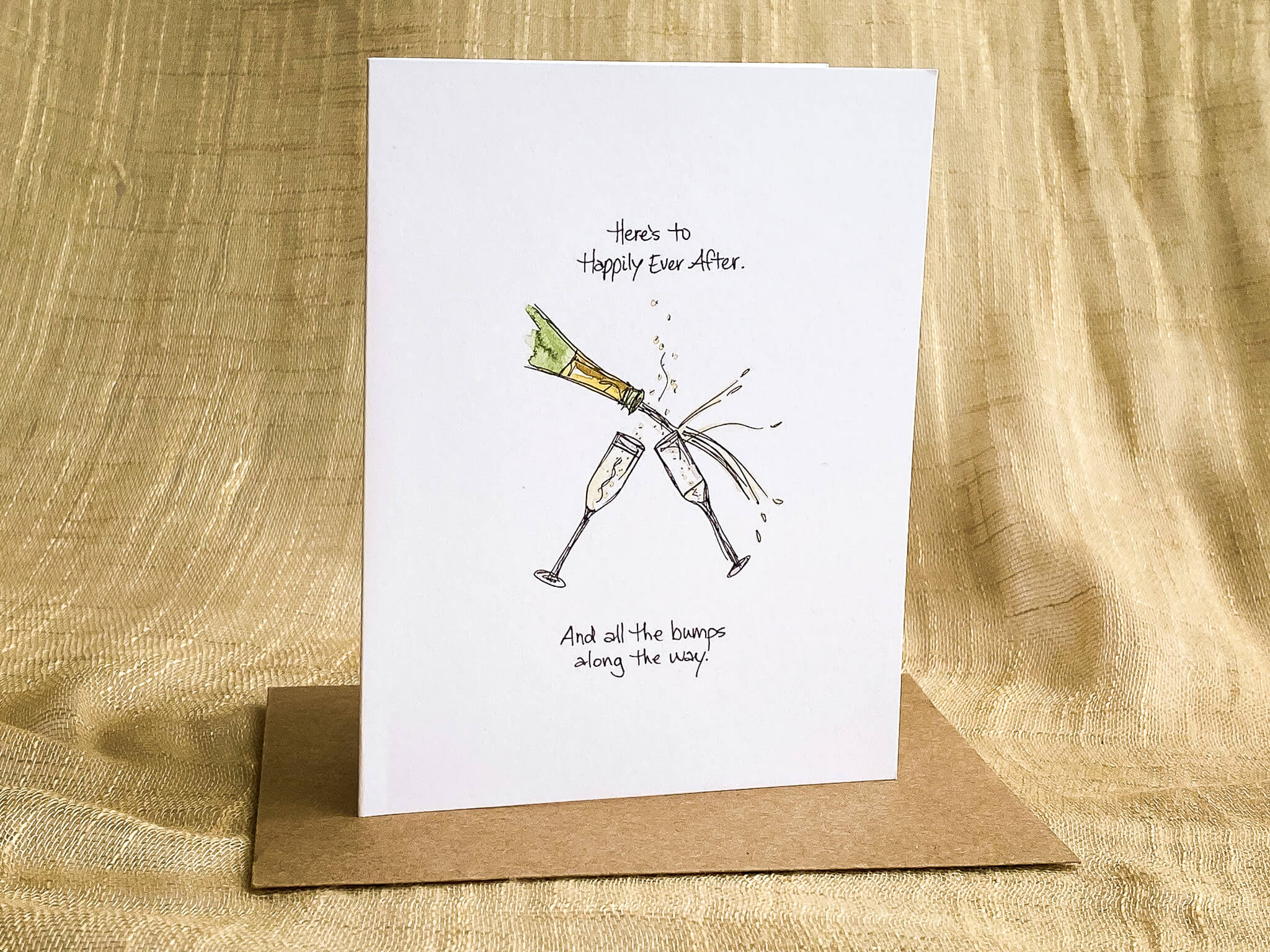 Wedding greeting card illustrated with clinking champagne glasses and the message 'Here's to Happily Ever After. And all the bumps along the way,' shown with a kraft paper envelope.