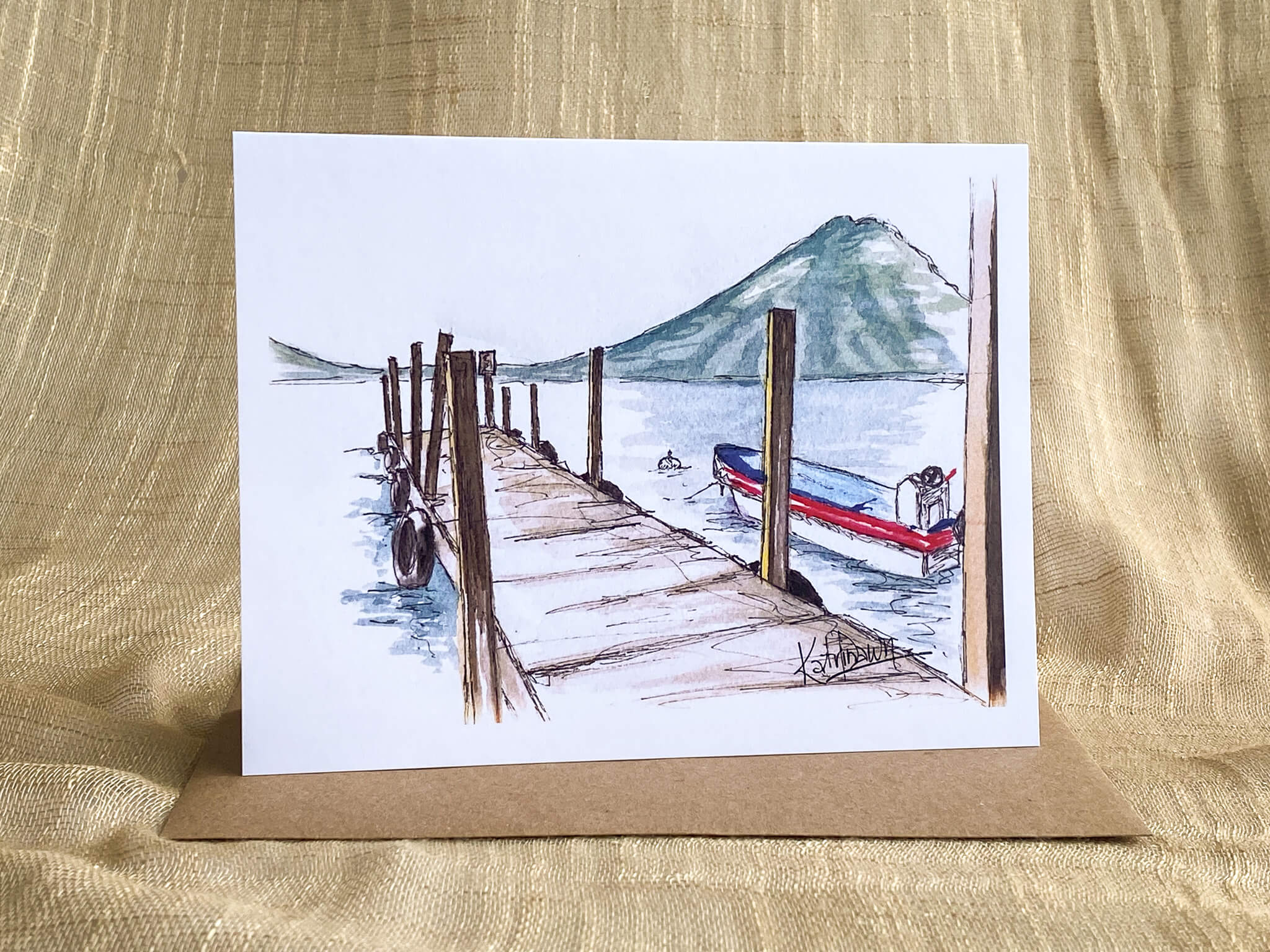 Watercolor greeting card by Katrina Dawn of Lake Atitlán, Guatemala, featuring a wooden dock, moored boat, and volcano backdrop, displayed with a kraft paper envelope.