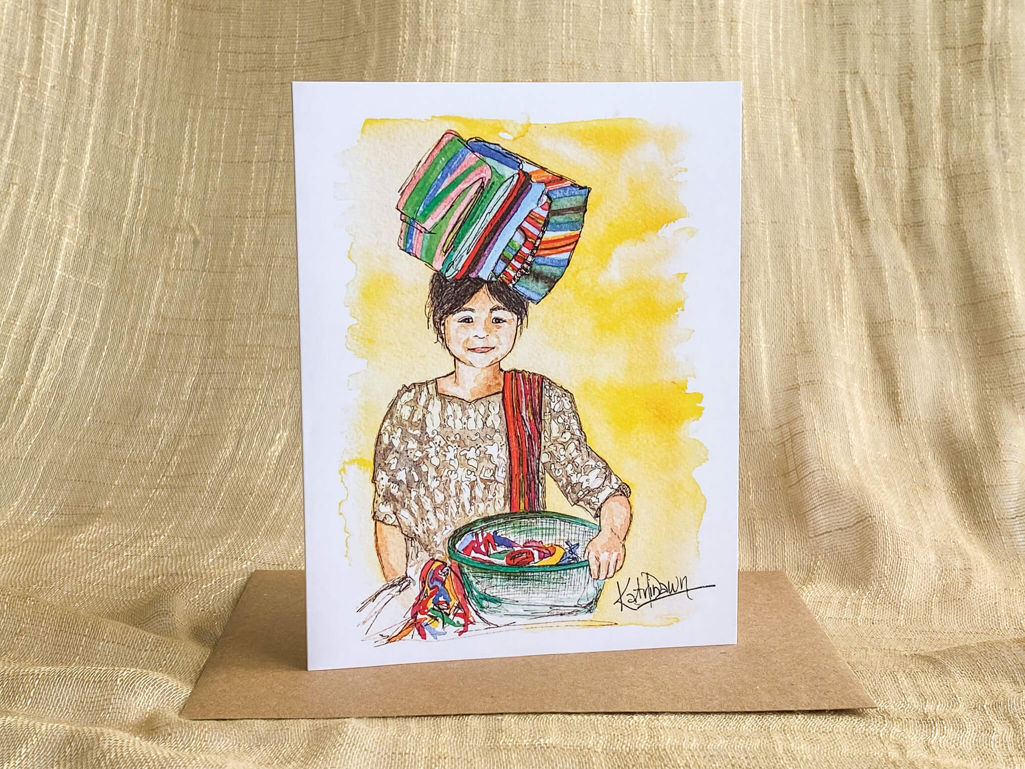 Watercolor and ink portrait greeting card by Katrina Dawn of a Guatemalan girl in traditional dress carrying a woven textile bundle on her head, set against a yellow watercolor background with a kraft paper envelope.