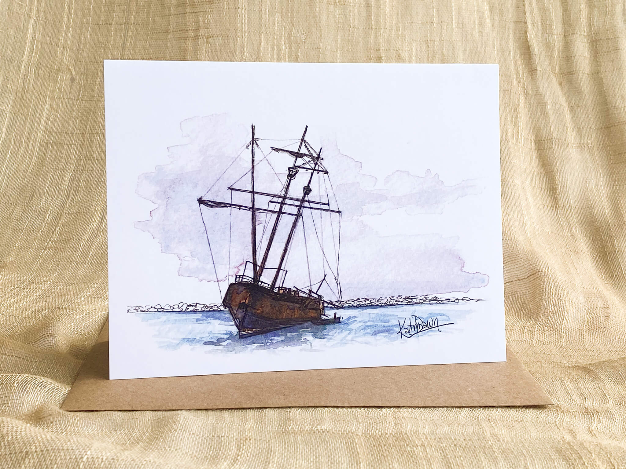 Watercolor sketch greeting card by Katrina Dawn of a weathered shipwreck with bare masts run aground on a rocky shoreline, shown with a kraft paper envelope.