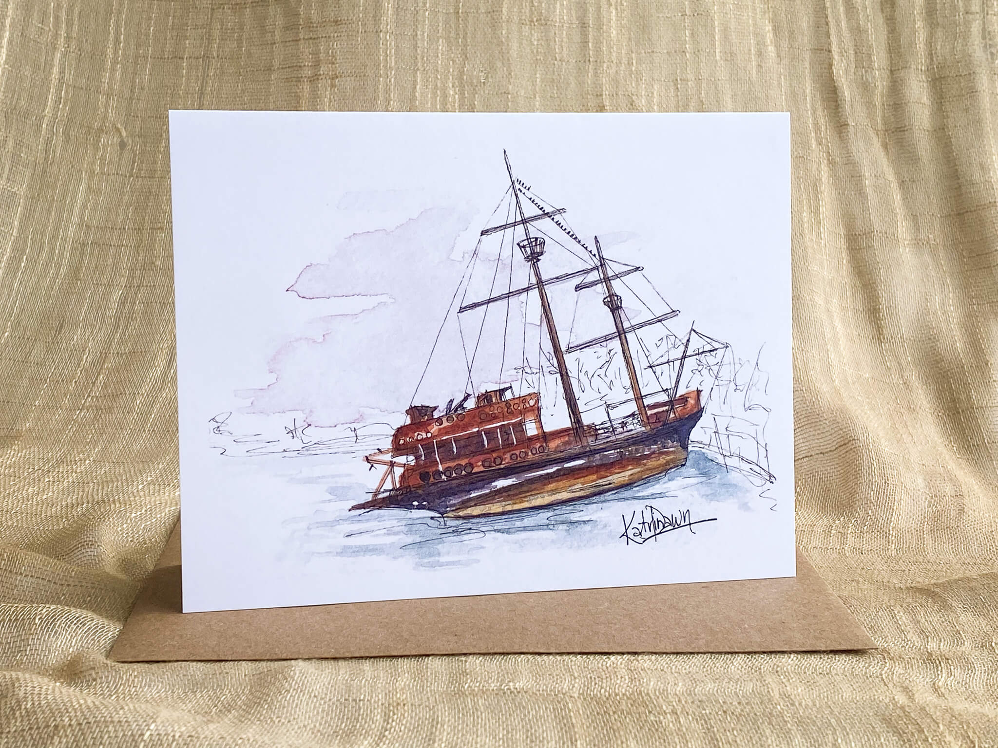Watercolor and pen greeting card by Katrina Dawn of a rustic shipwreck with tall masts and rigging resting in shallow water, shown with a kraft paper envelope.