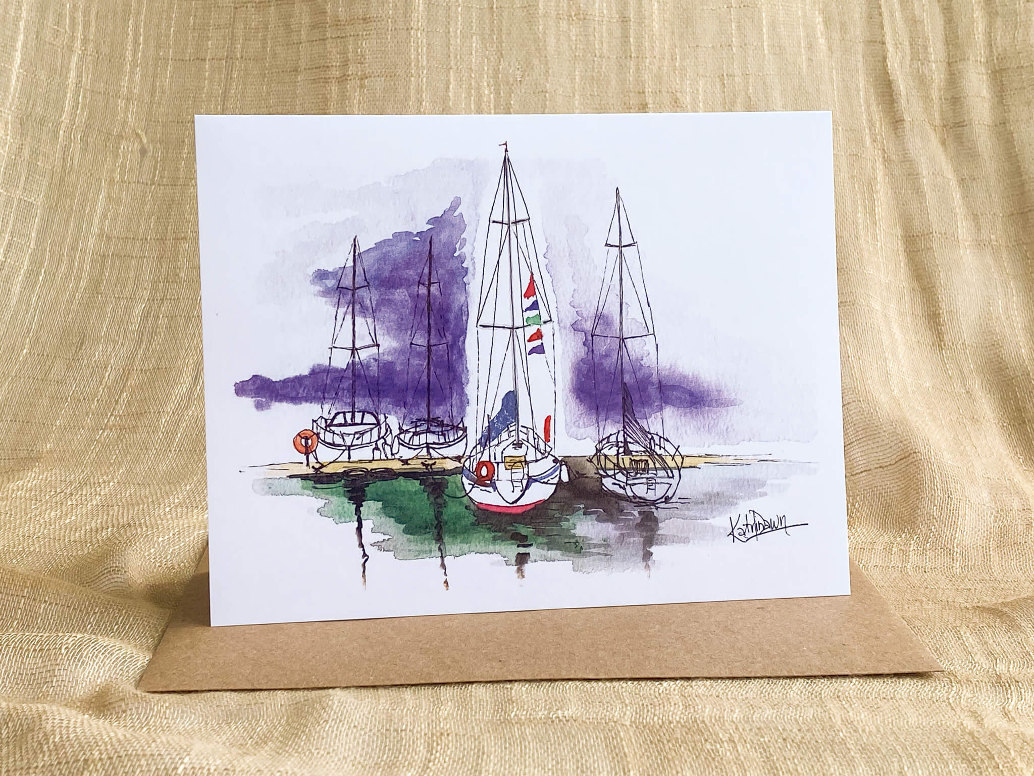 Watercolor and pen greeting card by Katrina Dawn of sailboats docked in a marina with purple watercolor sky and reflections, shown with a kraft paper envelope.