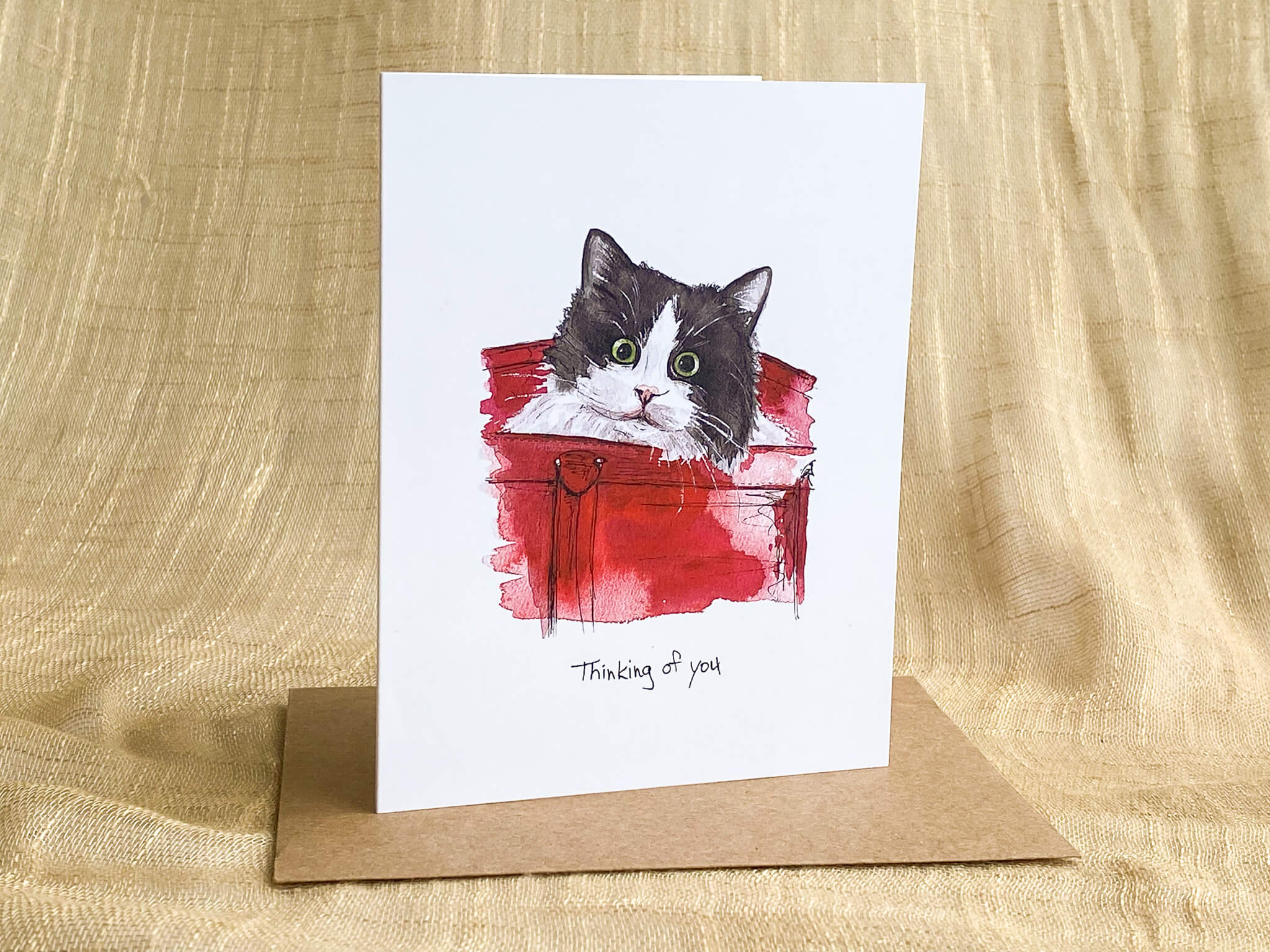 Watercolor greeting card by Katrina Dawn of a black and white cat peeking out of a red purse, with 'Thinking of you' script, paired with a kraft paper envelope.