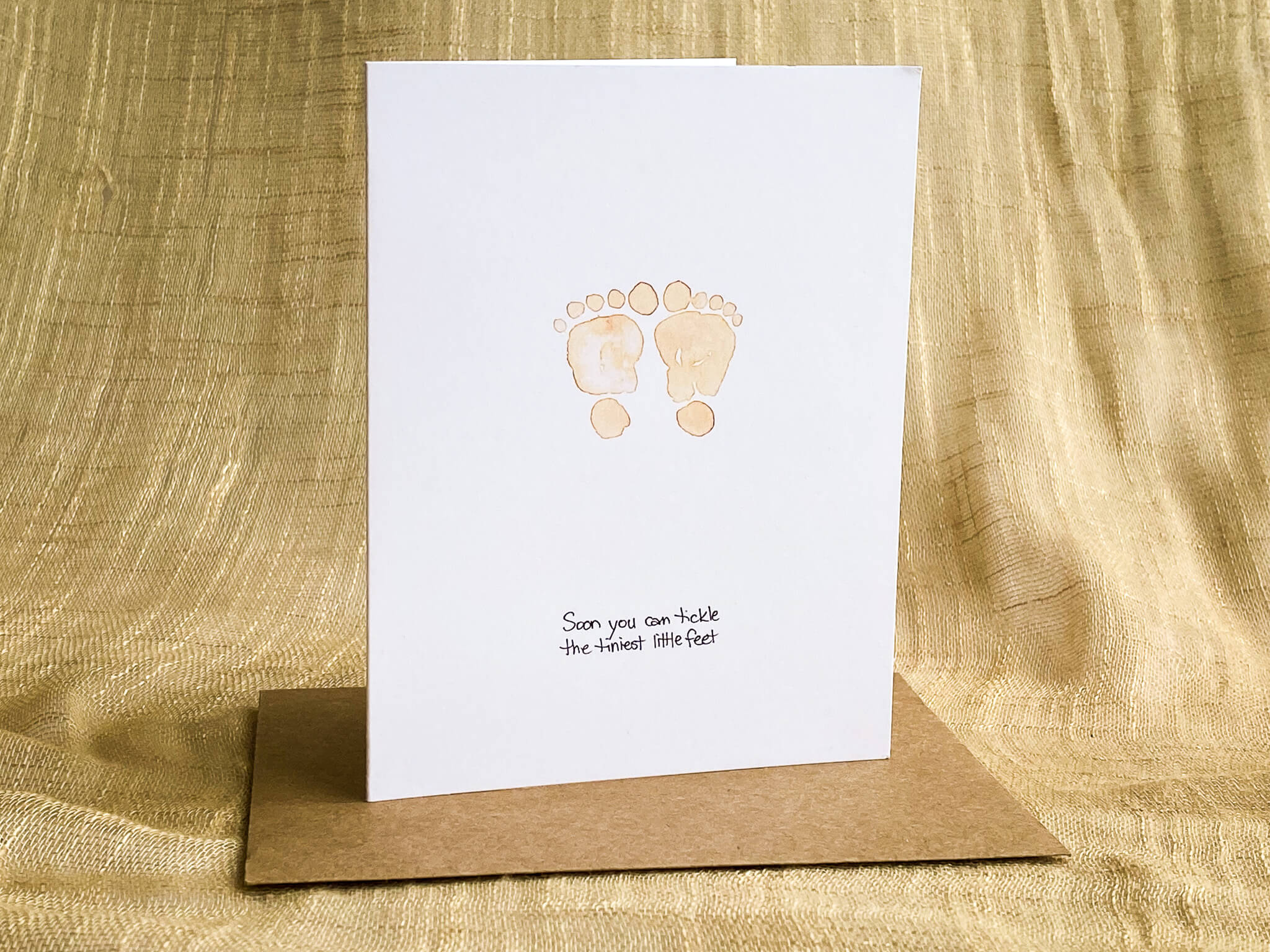 New baby greeting card by Katrina Dawn featuring hand-painted watercolor baby footprints with the message 'Soon you can tickle the tiniest little feet,' shown with a kraft paper envelope.