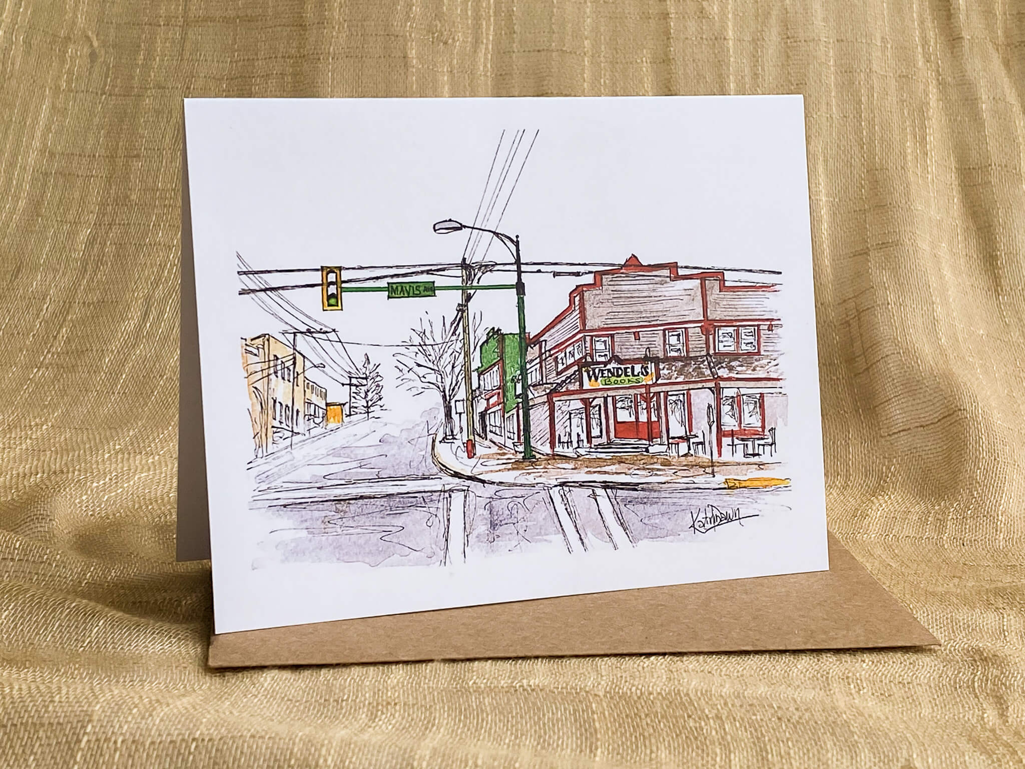 Watercolor and pen greeting card by Katrina Dawn of Wendel's Books storefront on a street corner in Fort Langley, with traffic signals and streetscape detail, shown with a kraft paper envelope.